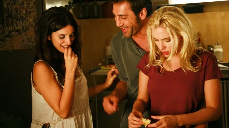 Tres personas jóvenes comparten un momento íntimo y distendido en una cocina, sonriendo y cocinando juntas, reflejando conexión y complicidad relacional.